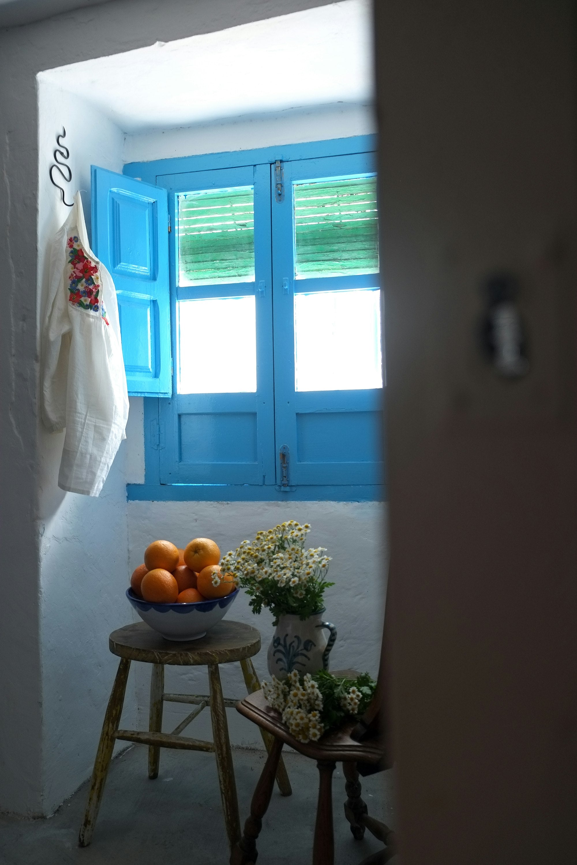 Rincón encalado con contraventanas azules, cuenco de naranjas y flores en jarra de cerámica